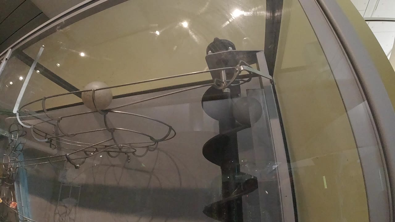 A curved glass display case fills the frame, with metal rods, circular wire loops, reflections, and dark shoe-like forms stacked near the right edge.