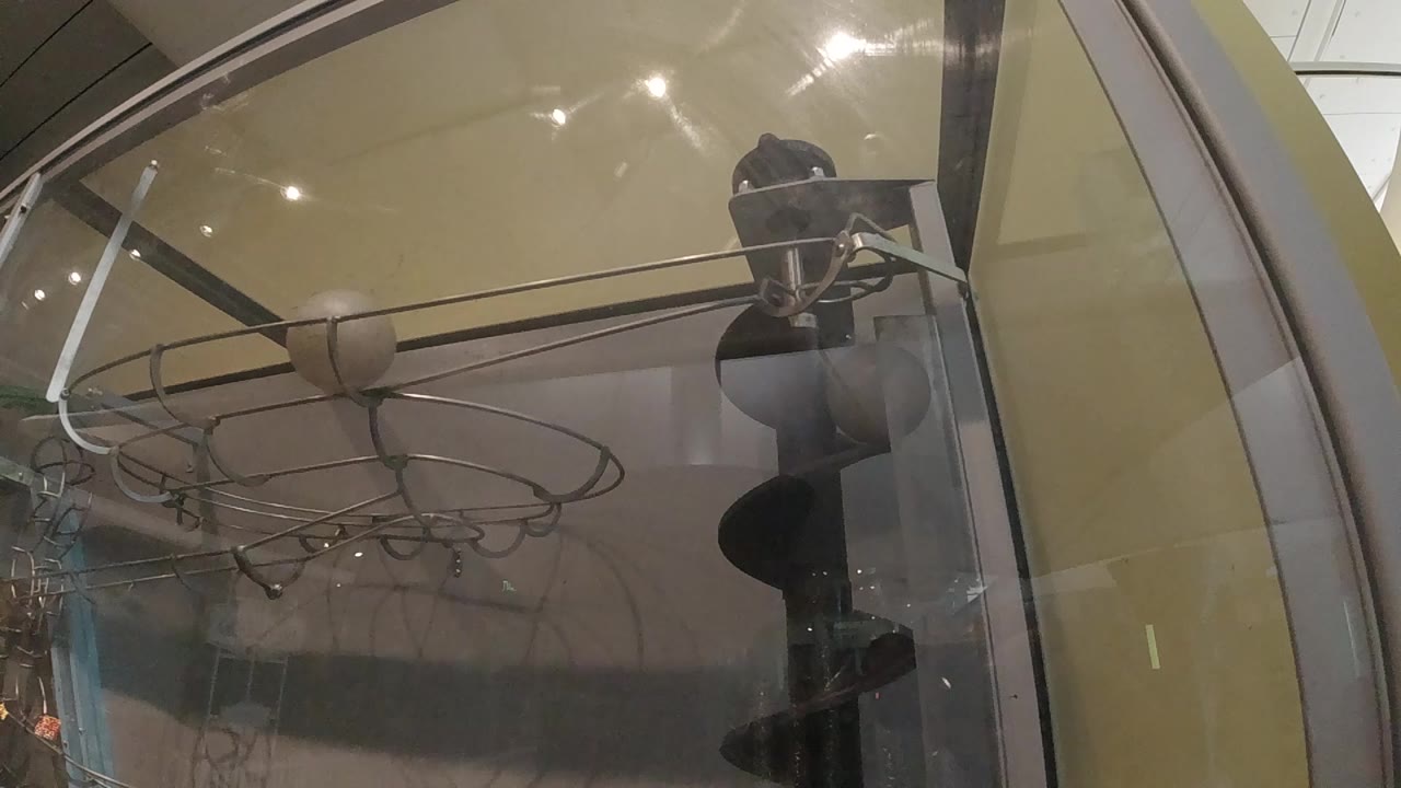 The glass display case remains close in frame, with a horizontal rod cutting across the upper area and dark shoe-like objects visible through reflections on the right.