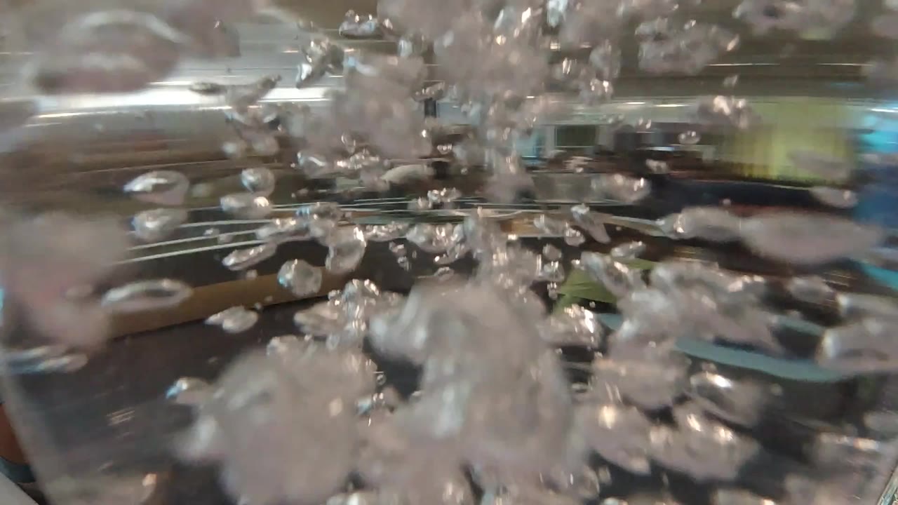 At 0:00, dense white air bubbles fill the center of a close-up liquid view in Denver, United States, with metallic reflections behind them and no visible people.