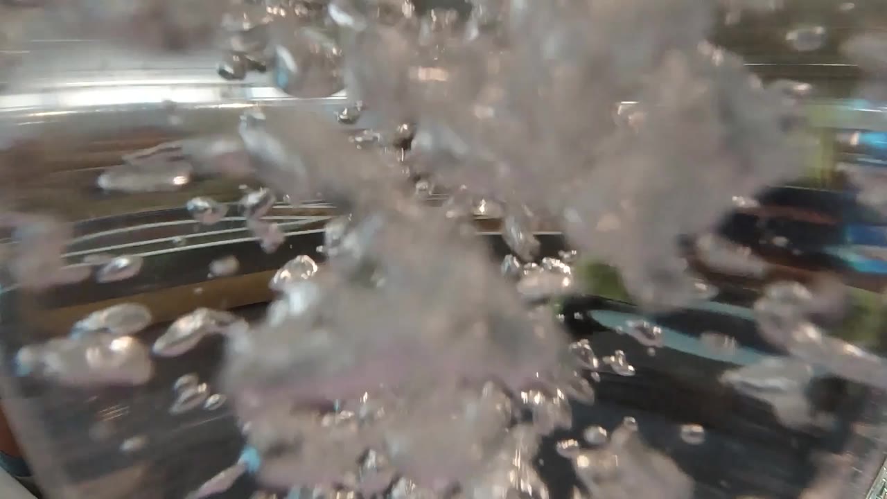 At 0:02, a cloudy vertical plume of bubbles rises through the center-right of the close-up, partially obscuring the reflective background and showing no visible people.