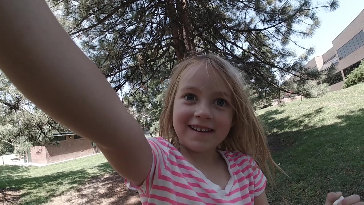 The girl smiles softly at the lens from a close selfie angle in a Denver park, with her forearm crossing the frame and a low building visible beyond the grass.
