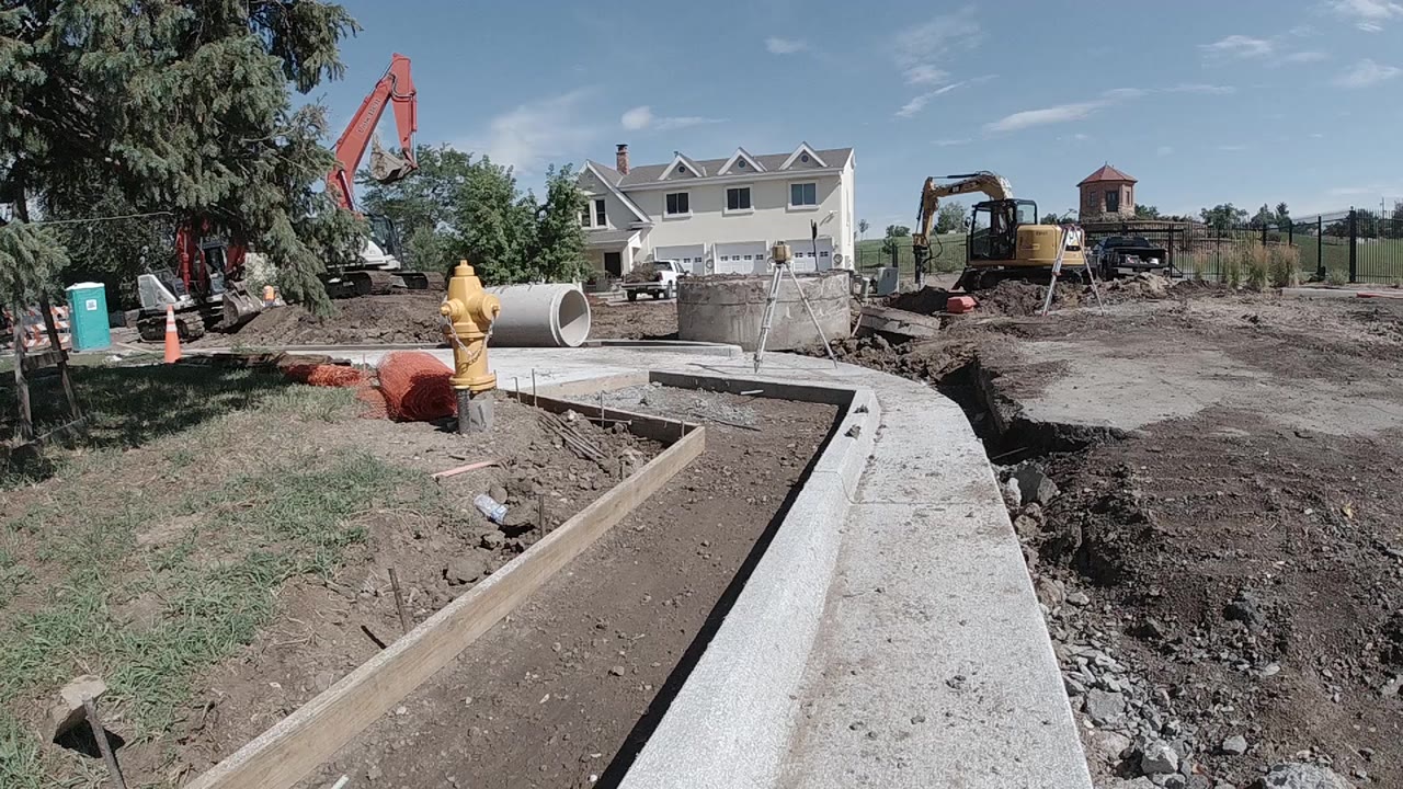 Aerial drone view of a Denver construction site showing a concrete-lined drainage channel across bare earth, red excavator arm at left, yellow machinery midground, partially built concrete block walls at right, and a treeline under blue sky; no visible people.