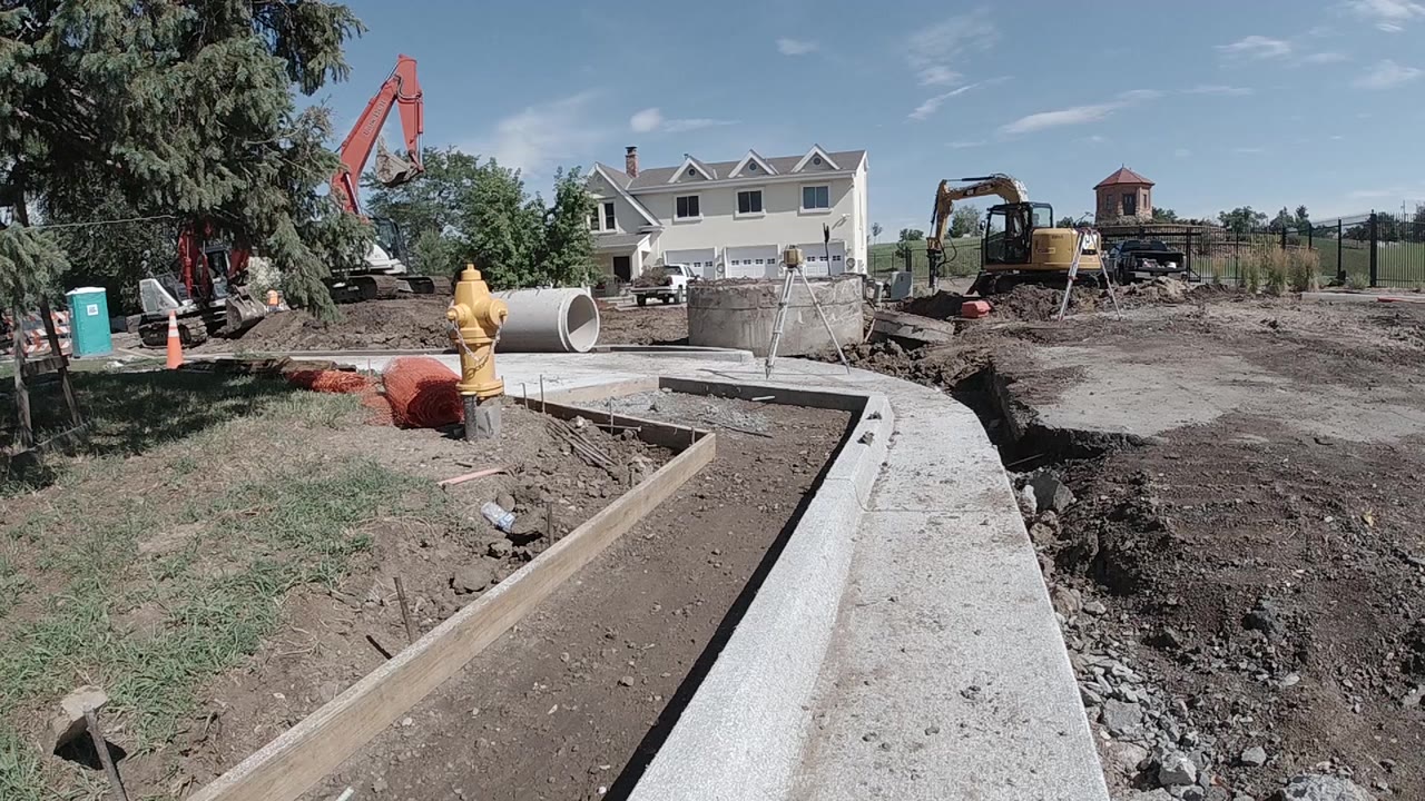 Aerial drone view of the Denver construction site showing the concrete drainage channel, red excavator arm at left, yellow machinery midground, partially built concrete block walls at right, and treeline; no visible people.