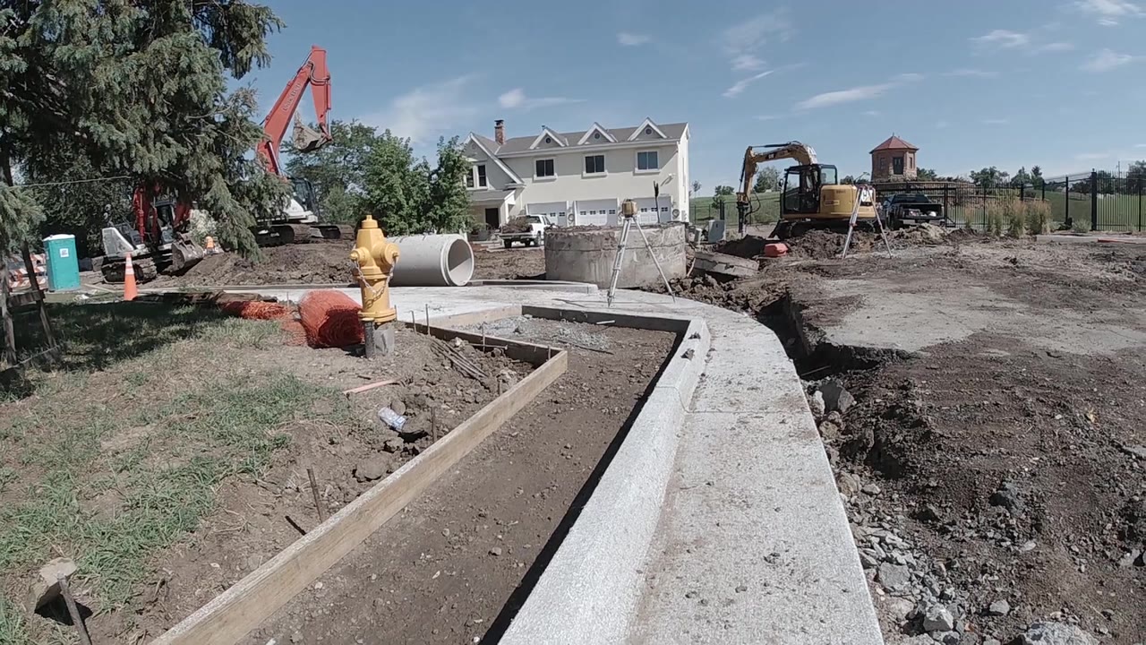 Aerial drone view of the Denver construction site at two seconds showing the concrete drainage channel, red excavator arm, yellow machinery, partially built concrete block walls, and treeline perimeter; no visible people.