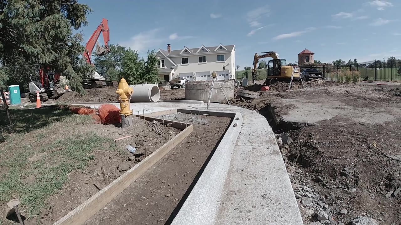 Aerial view of a construction site with concrete drainage channel, red excavator arm, yellow equipment, and orange safety markers in the midground.