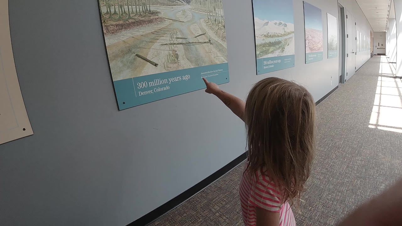 The child remains under the museum display in Denver with her arm extended toward the poster text and a bright hallway stretching into the background.