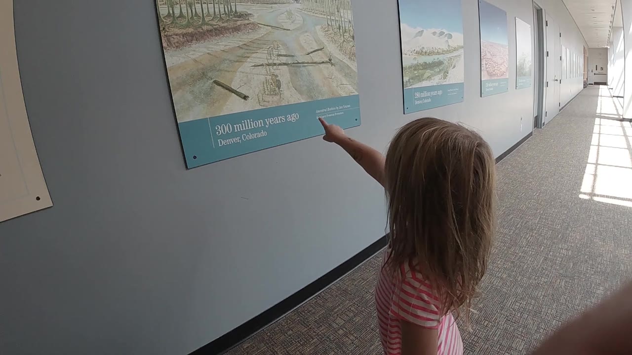 The blonde girl points at the lower portion of a museum poster in a Denver hallway, with more informational panels and sunlit windows visible down the corridor.