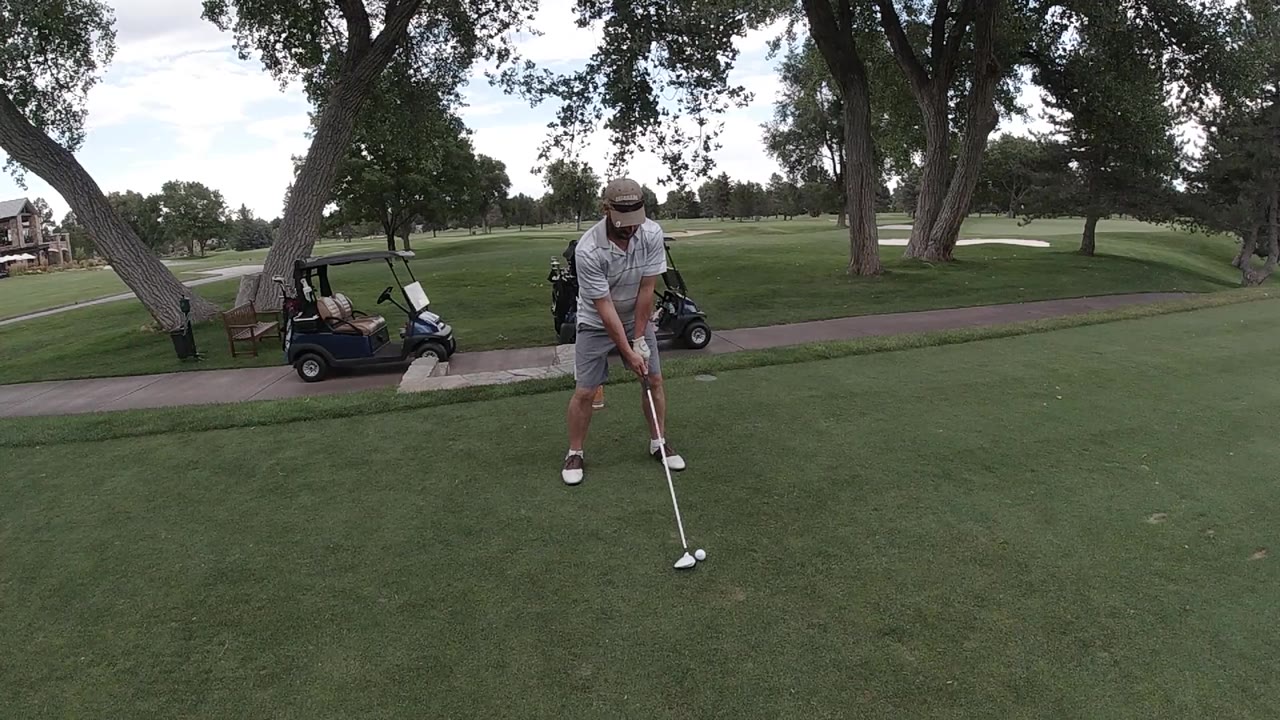 At 0:01, the Denver golfer holds his stance with the club near the ball while the cart path and parked golf cart sit behind the tee area.