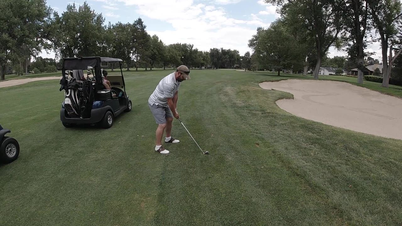 Golfer addresses a ball beside a golf cart on a green fairway.