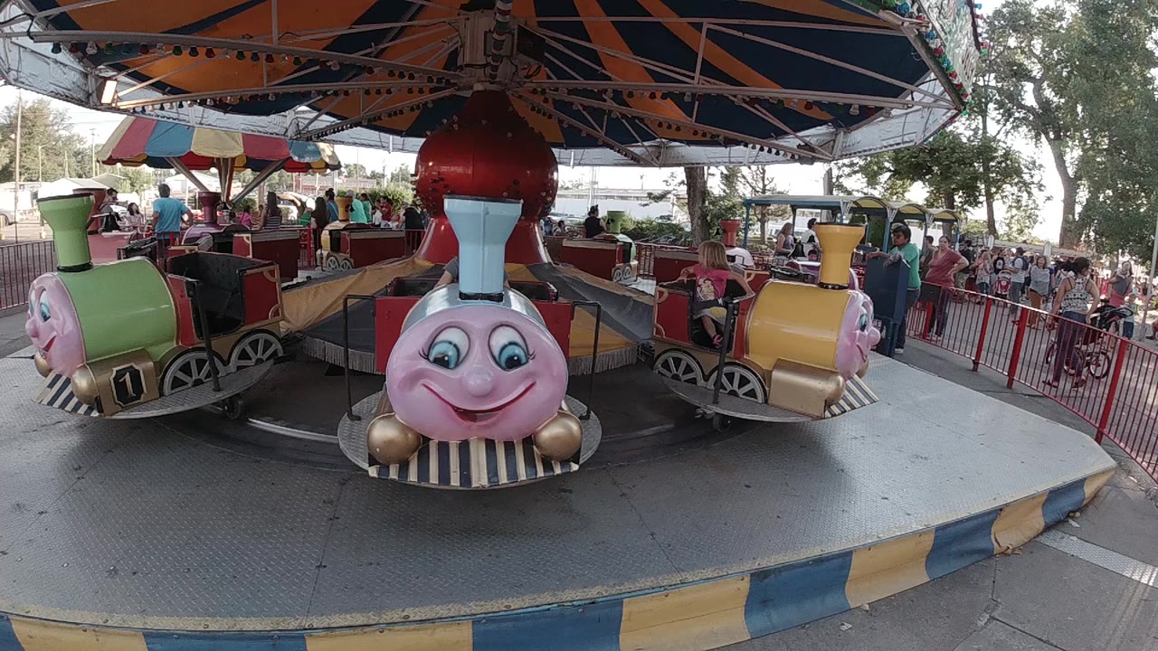 A blue and pink smiling train car faces the camera on a small circular ride in Denver, with red railings, seated riders, and trees behind it.