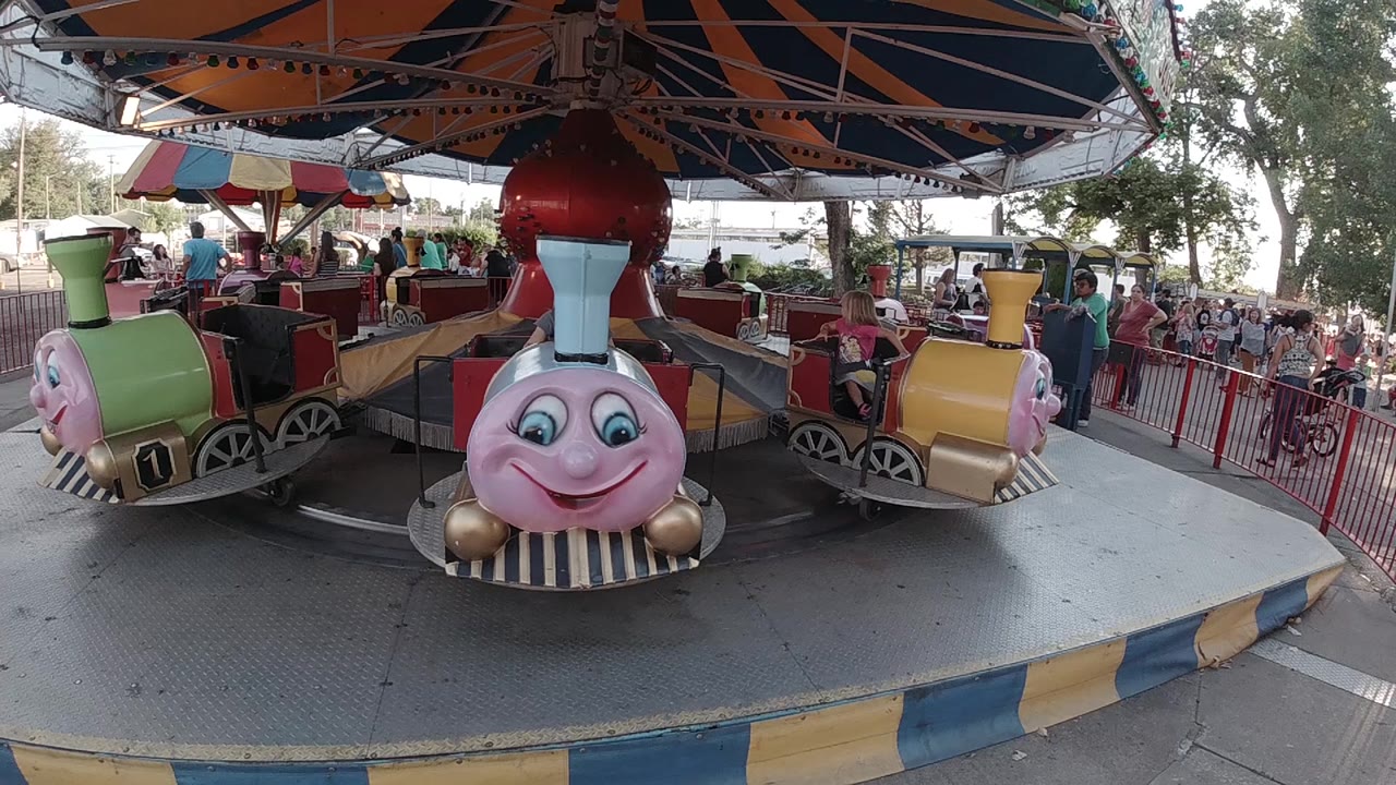 The smiling train car remains near center under the orange and dark striped canopy, with a yellow car and people waiting behind the red fence.