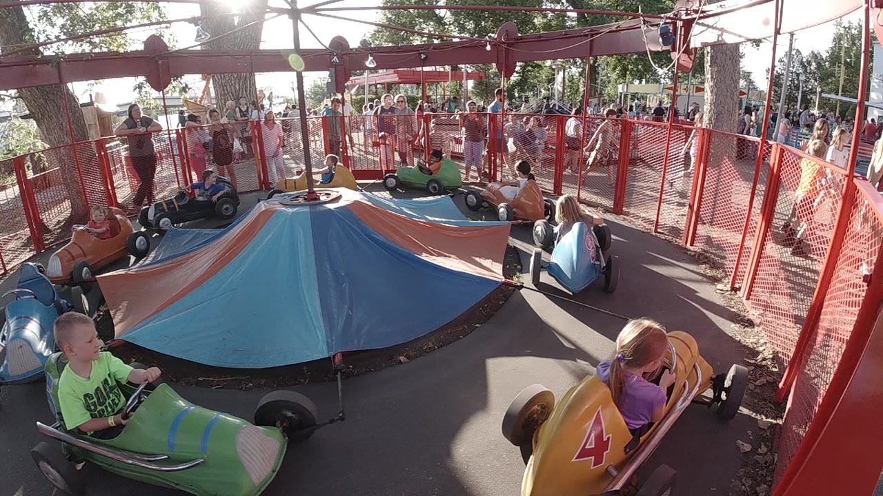 At Denver's Lakeside Amusement Park, children in green and yellow cars circle a blue and orange canopy with red fencing and spectators behind them.