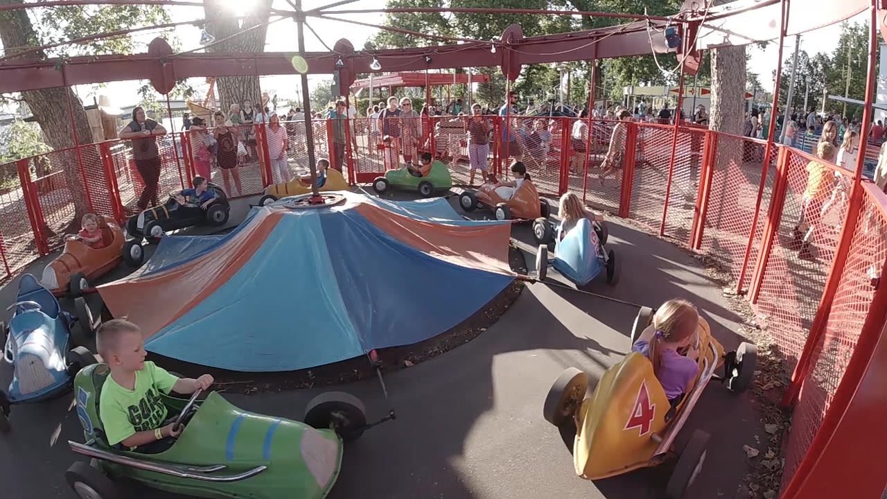 The yellow car with a child driver turns along the right side as a green car and several blue cars remain visible around the canopy at the Denver amusement park.