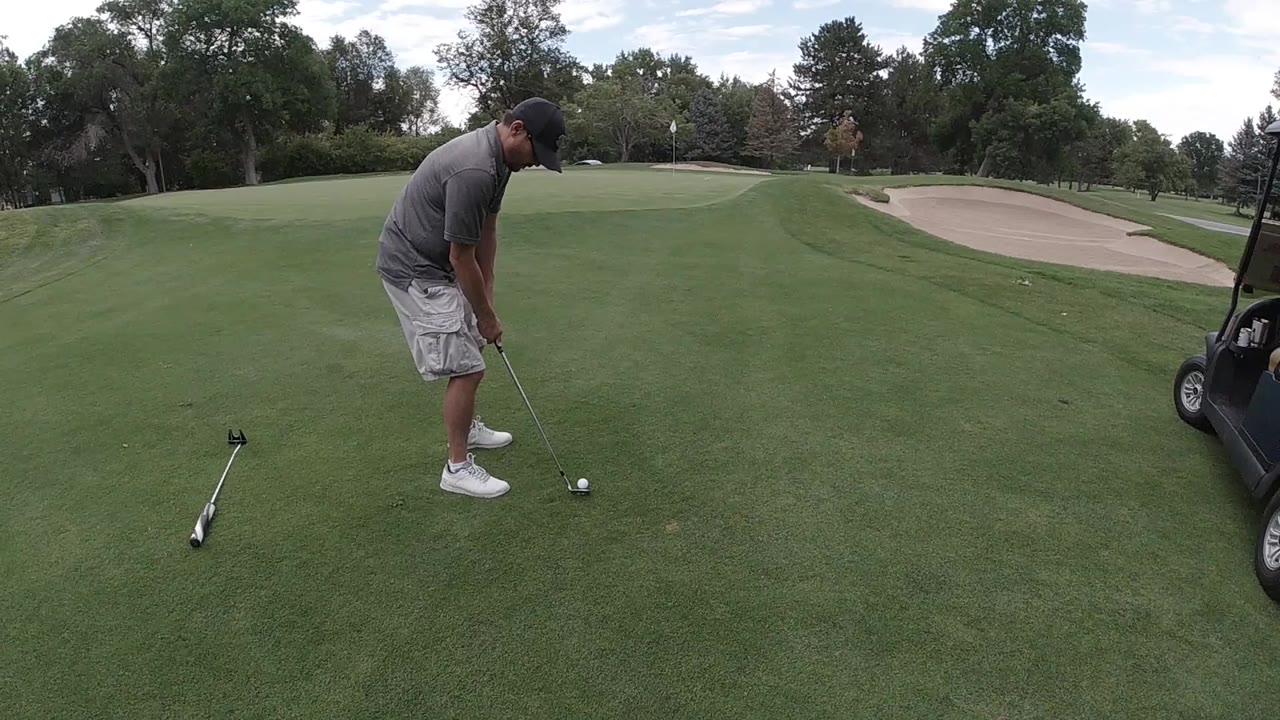 In Colorado, United States, an adult golfer bends over a ball on a manicured fairway while a spare club lies on the grass and a bunker sits beyond him.