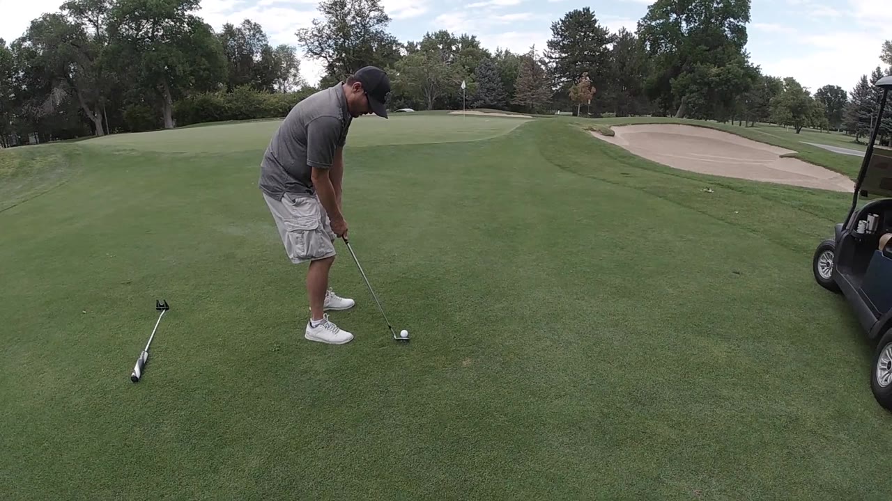 An adult golfer in a gray shirt and white shoes lines up a shot on the fairway, with the sand bunker and tree line still fixed in the background.