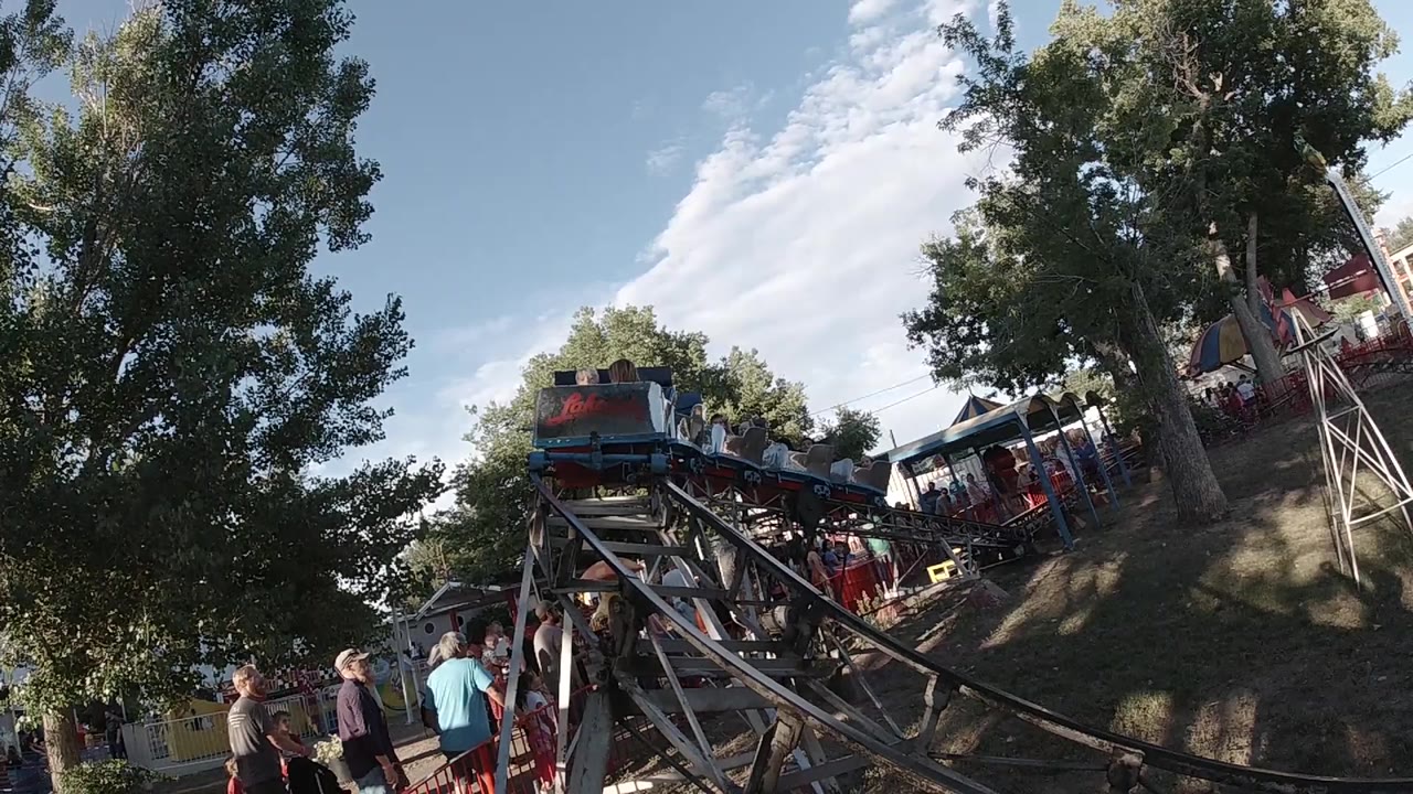 A blue and red kiddie rollercoaster approaches from the background on a dark track in Colorado, United States, with trees, sky, spectators, and riders visible.