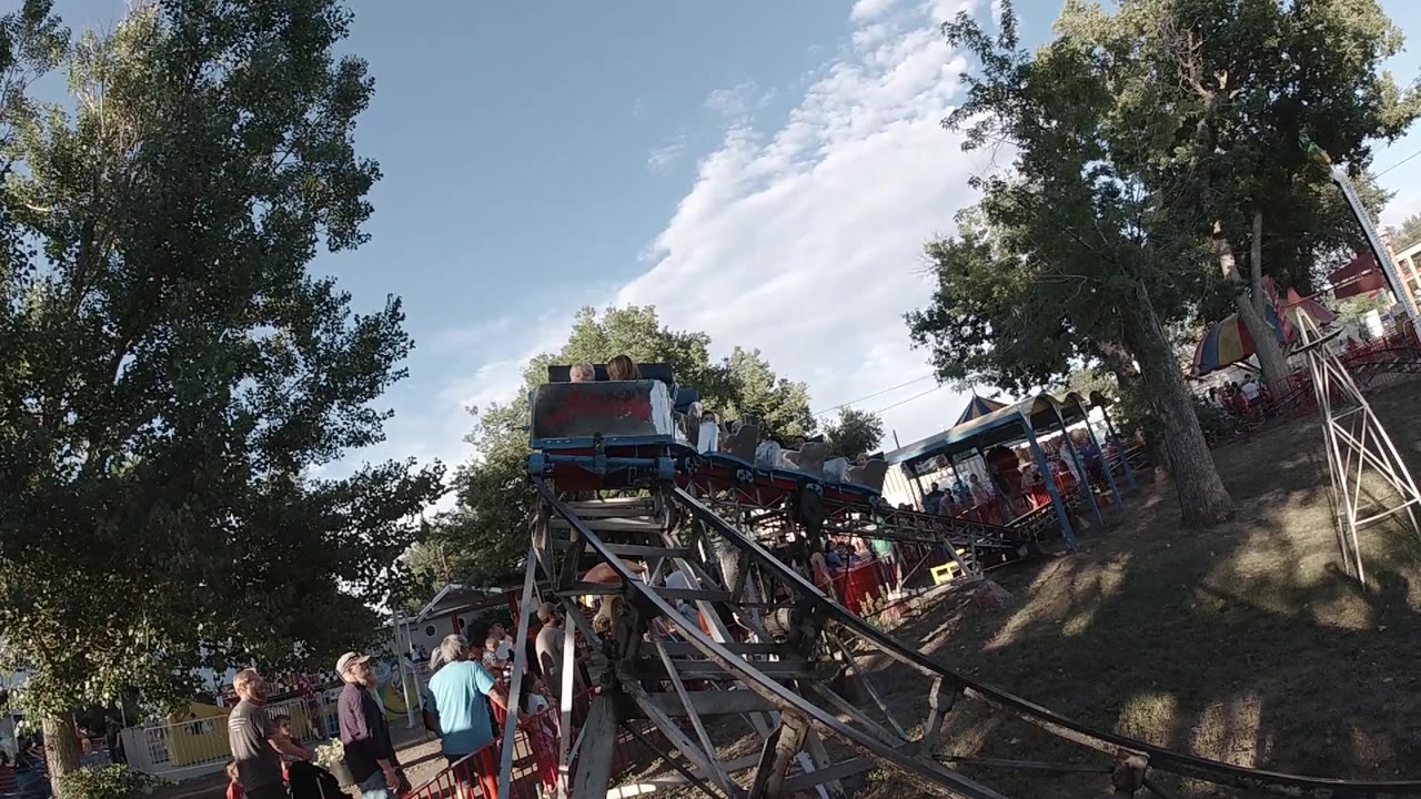 A small coaster car advances along the rails under tall trees, with visible riders, spectators beside the track, and a bright sky over the Colorado carnival setting.