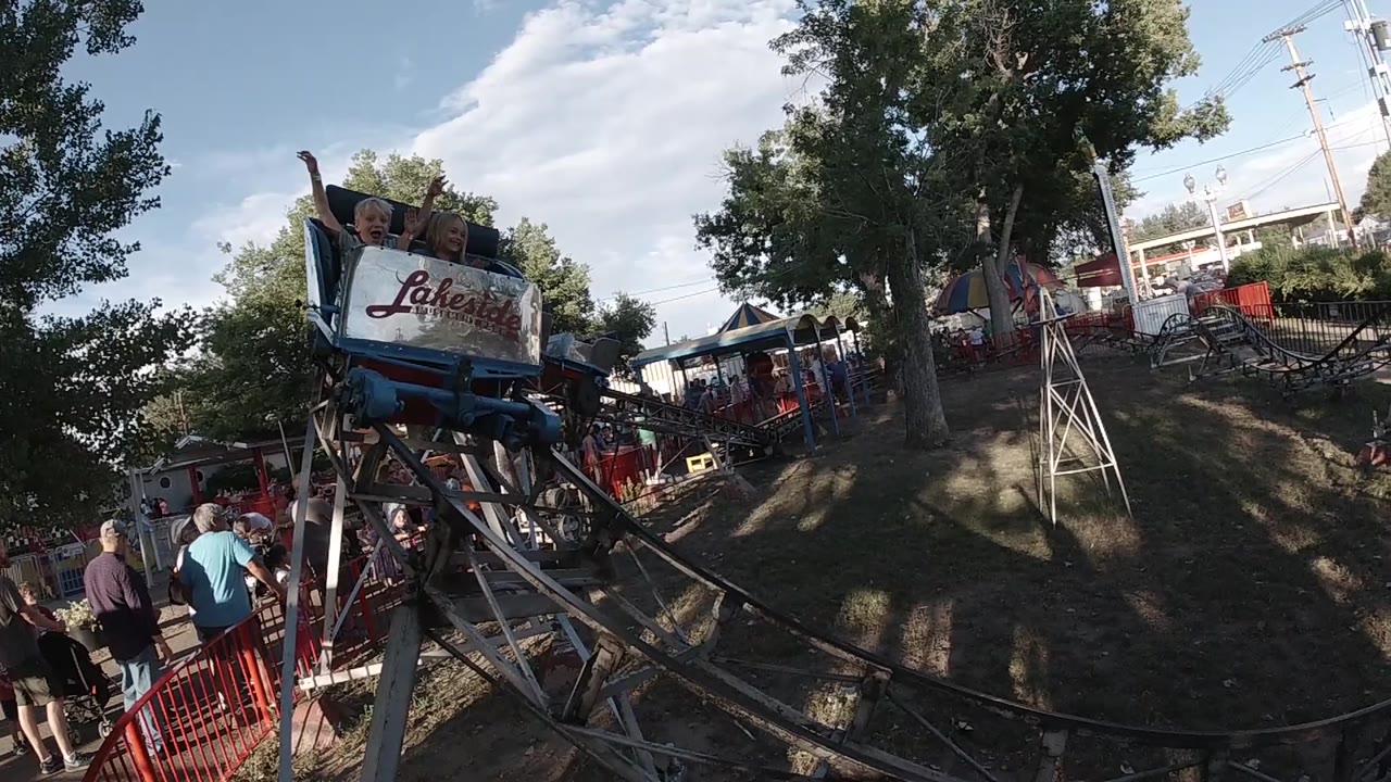 Children sit in the front of a small blue and red coaster car in Colorado, with the track curving past trees and fairground structures in the background.