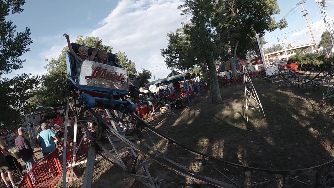 The kiddie coaster car moves closer through the frame, showing children seated under tree shade with black track rails sweeping across the foreground.