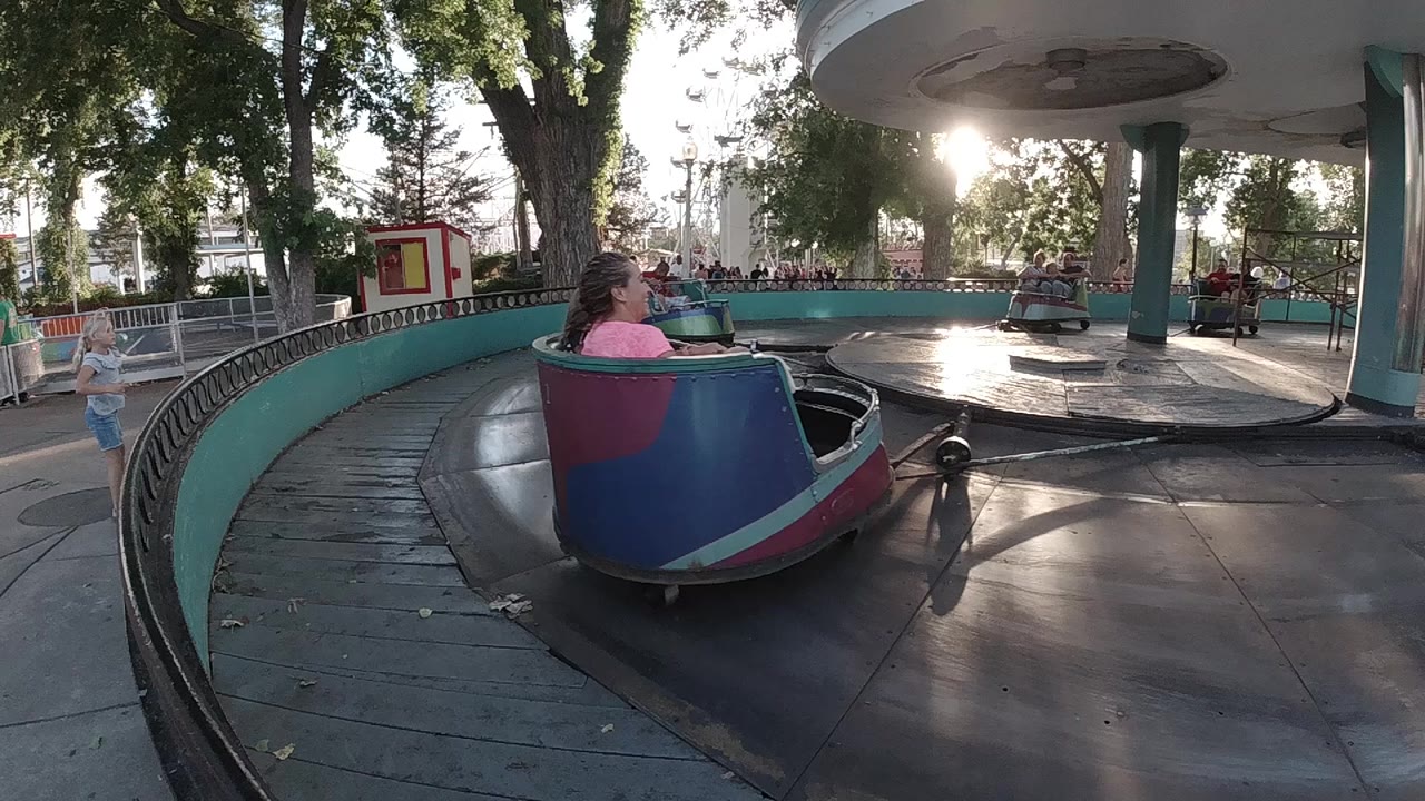 A colorful cup ride car with a child in a pink shirt passes near the camera on a circular amusement park platform in Colorado.