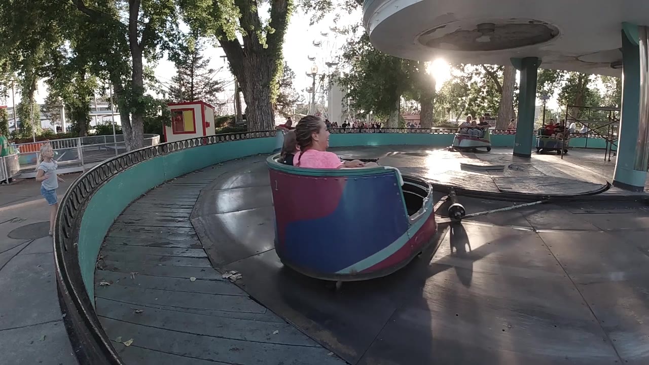 A blue and magenta cup car reaches the right side of the frame while the ride platform, canopy column, and background fence remain visible.