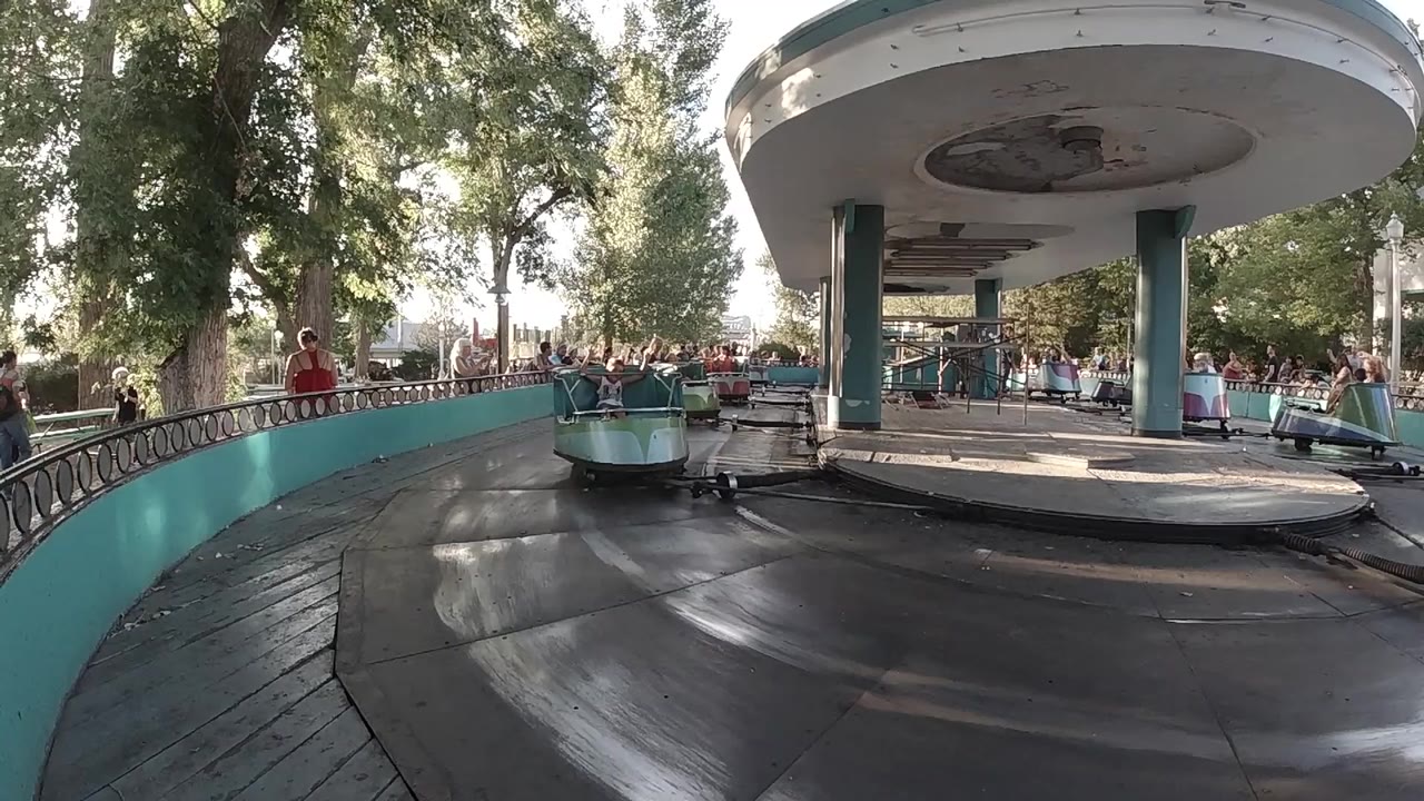 Spinning ride cars under trees beside a teal barrier.