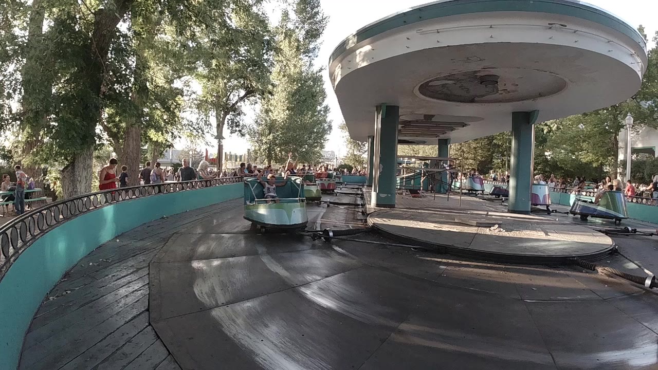 A low view of a colorful carousel ride in Colorado shows a teal car on a dark circular track, with park visitors behind railings and trees in dappled light.