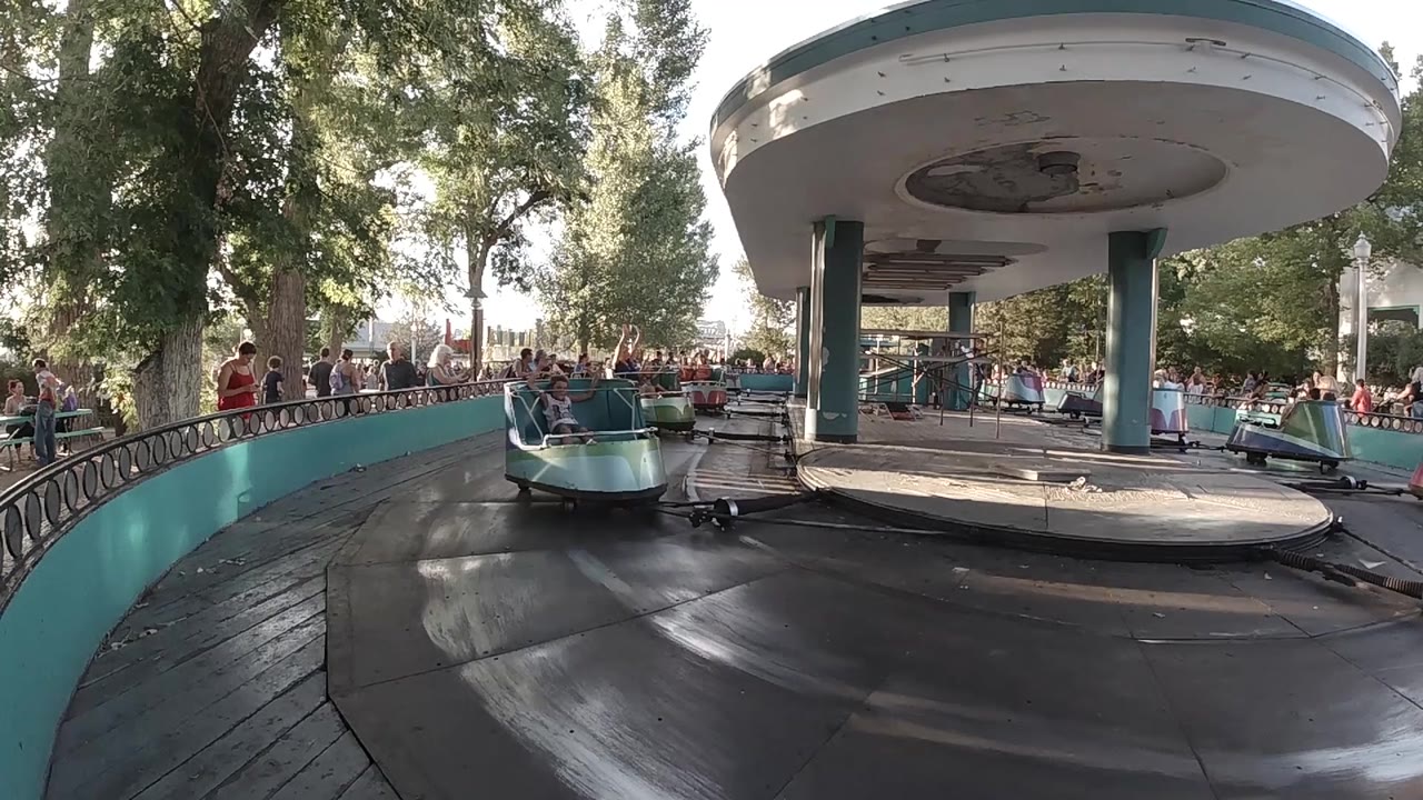 A teal and blue carousel car passes beside the curved teal barrier, with a busy line of visitors, trees, and the canopy structure in the background.