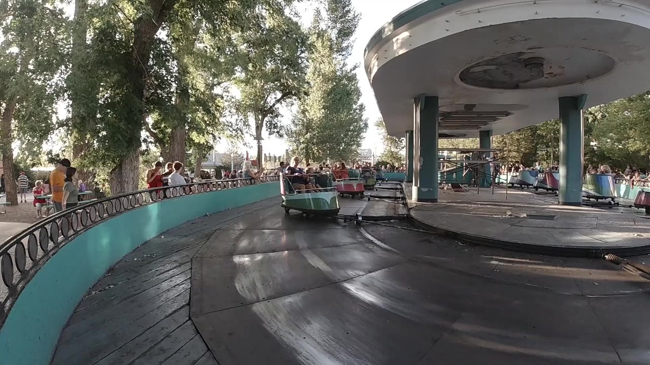A low view at Denver's Lakeside Park shows a teal and magenta ride car ahead on a dark circular track with visitors behind railings.