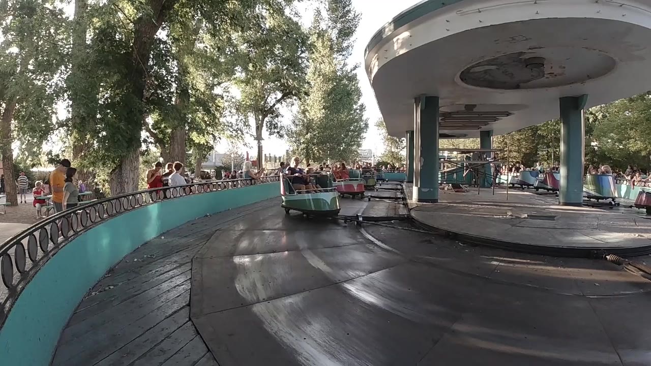 A teal ride car moves closer on the circular track while a turquoise wall, trees, and a crowded railing frame the Denver amusement park scene.