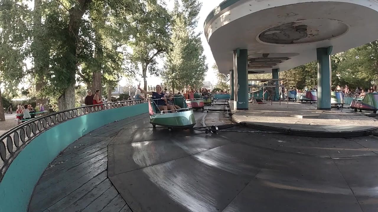 A cup carousel car approaches from the left in a Colorado amusement park scene, with riders visible under trees and a round canopy ahead.