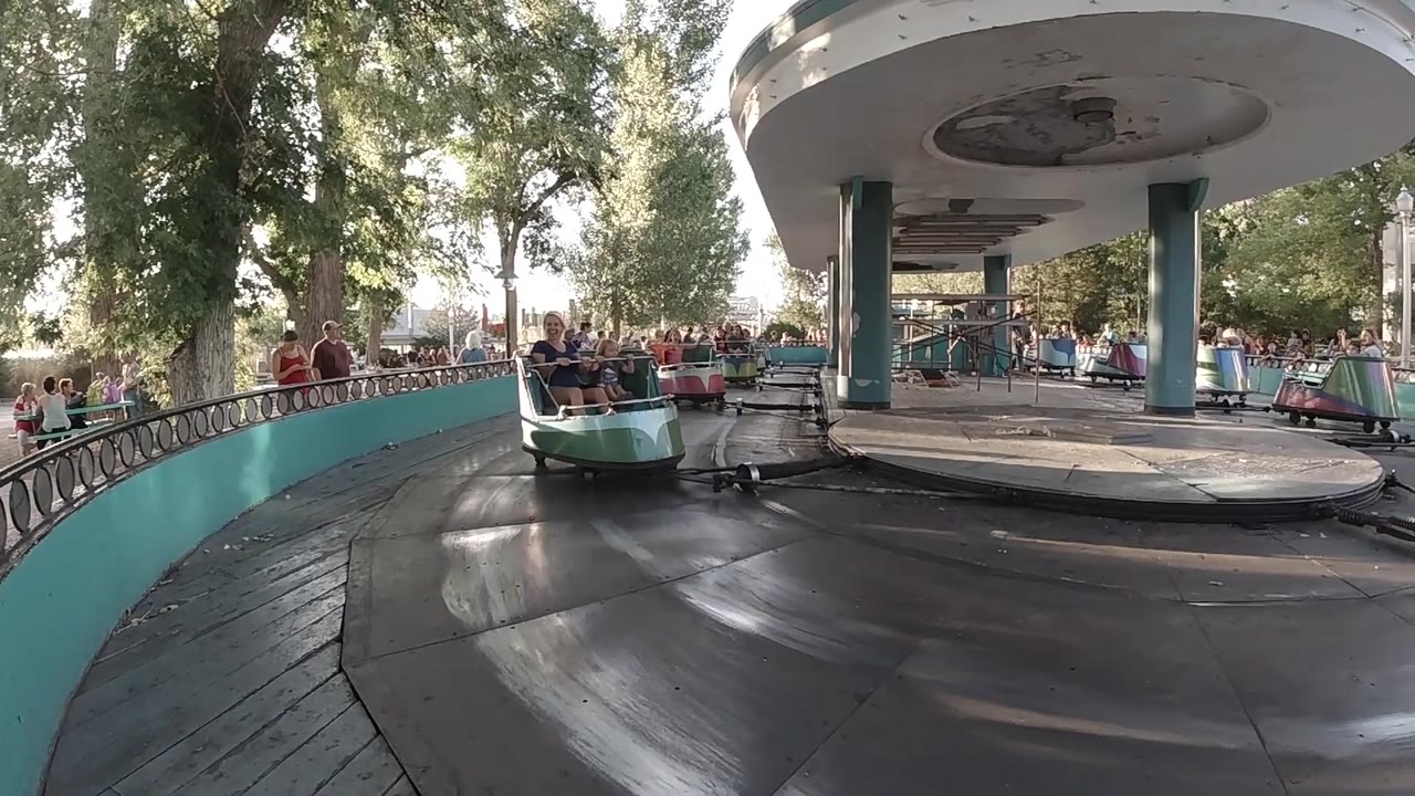 A cup carousel car passes in front of the white canopy and teal posts, with people riding, people watching, and trees filling the Colorado park background.