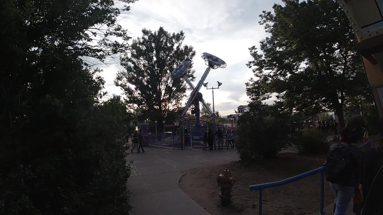 The ride structure sits behind a central tree, with visitors clustered near the gate and a blue handrail along the right foreground.