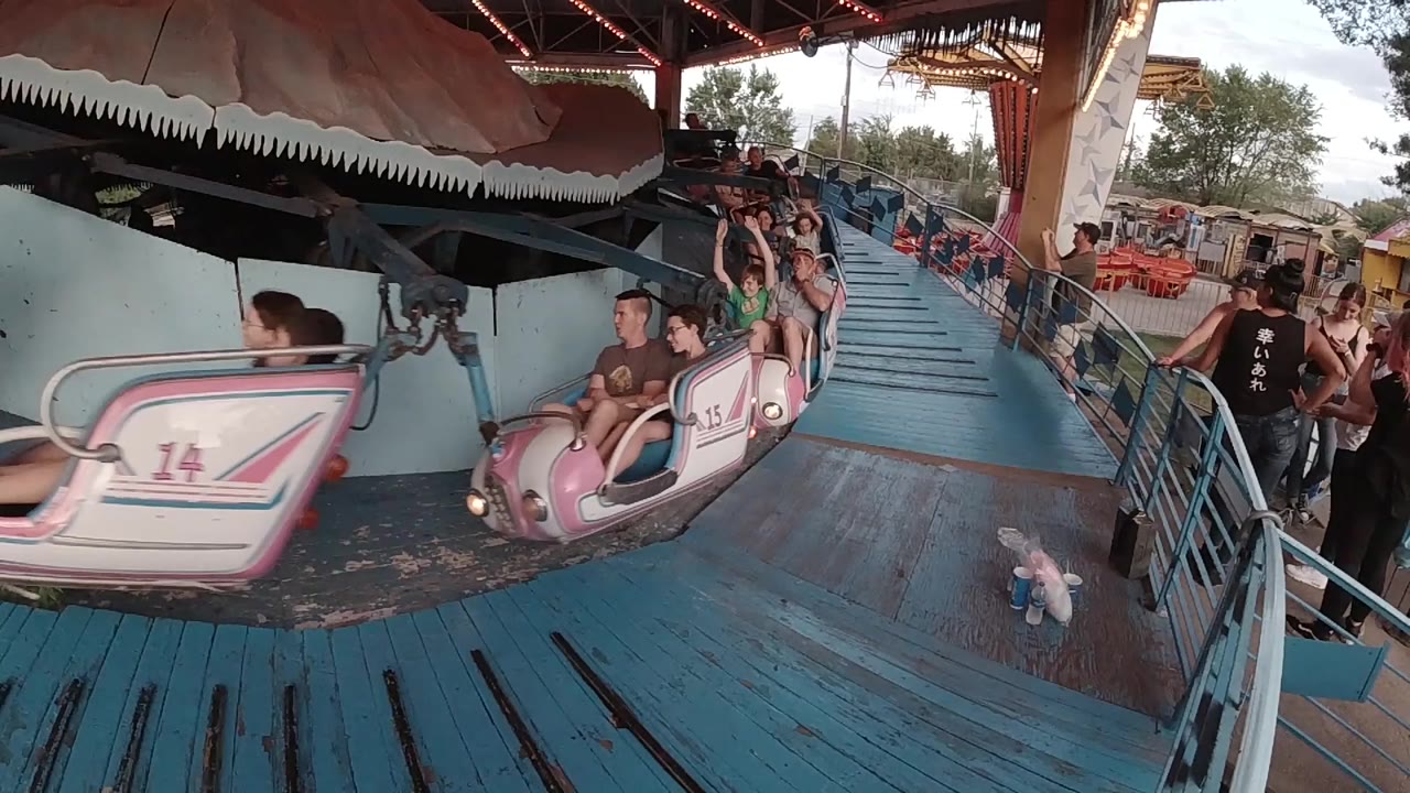 At 0:00 in Denver, pink spinning ride cars move past a blue wooden platform while riders and waiting guests are visible under the covered structure.