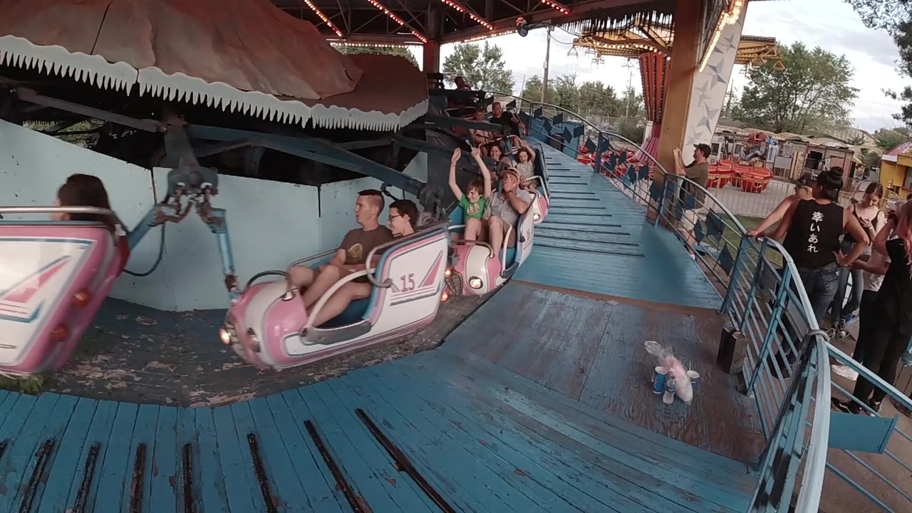 At 0:02, the left ride car partially exits frame while another pink car with riders approaches beside the blue platform in daylight.