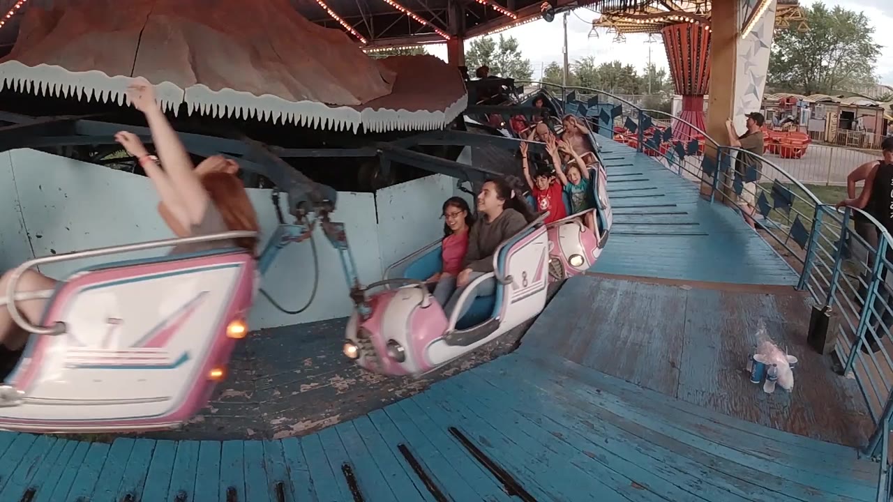 Pink Music Express cars pass close to the camera on a blue platform in Denver, with children and adults visible under the ride canopy.