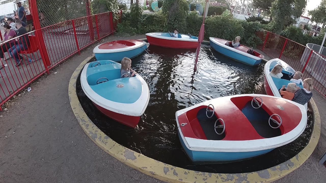 Colorful speedboat ride cars float around the water channel, with the nearest blue boat at left and a boat carrying adults near the right railing.