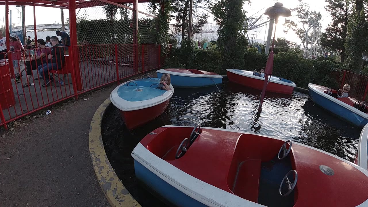 A Colorado amusement park speed boat carousel shows red, white, and blue boats circling dark water while children ride and families wait behind red railings at the left.
