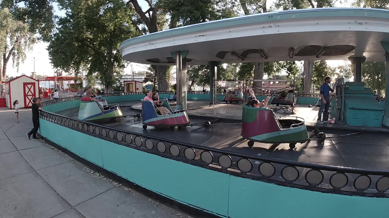 At 0:01, riders remain visible on the circular boat carousel while an adult stands near the right side beside a teal control booth or ride structure.