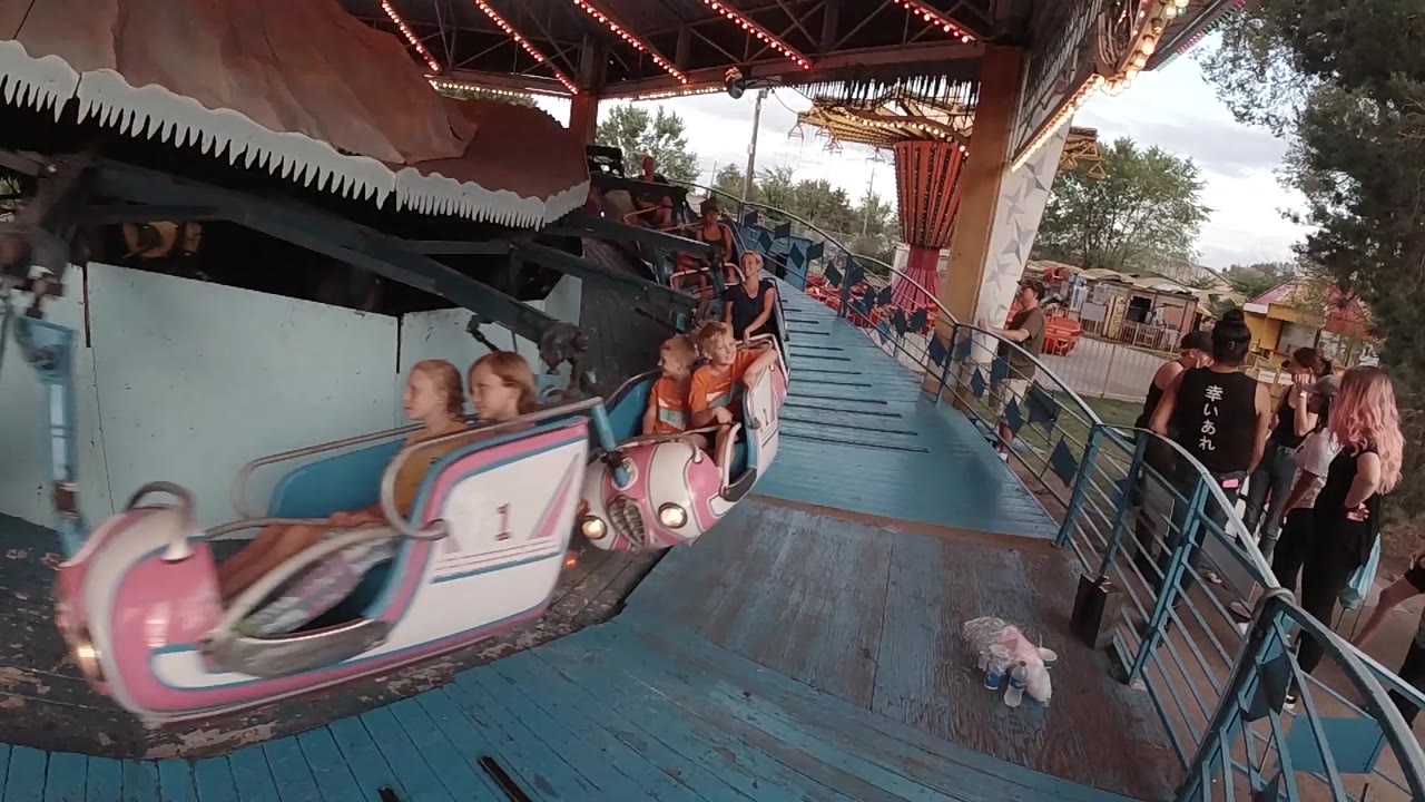 A pink numbered amusement ride car passes close to the camera beside a blue loading ramp in Colorado, with riders and waiting adults visible.
