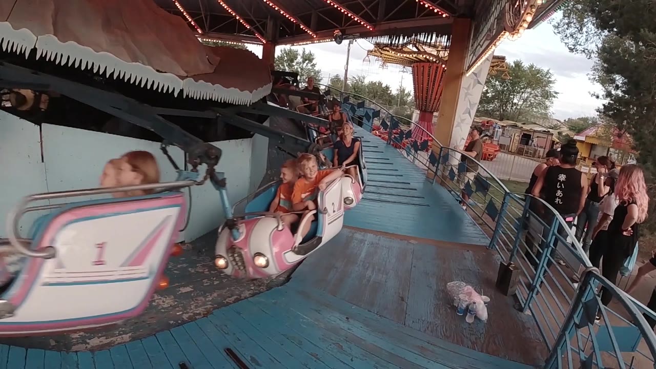 A numbered pink and white ride car slides across the foreground in Colorado, with seated passengers and a busy loading rail behind it.