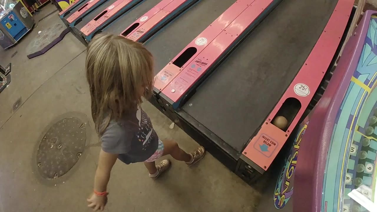 A young blonde girl in Colorado stands near the start of pink skee ball lanes, with the ramp and a circular arcade game edge visible beside her.