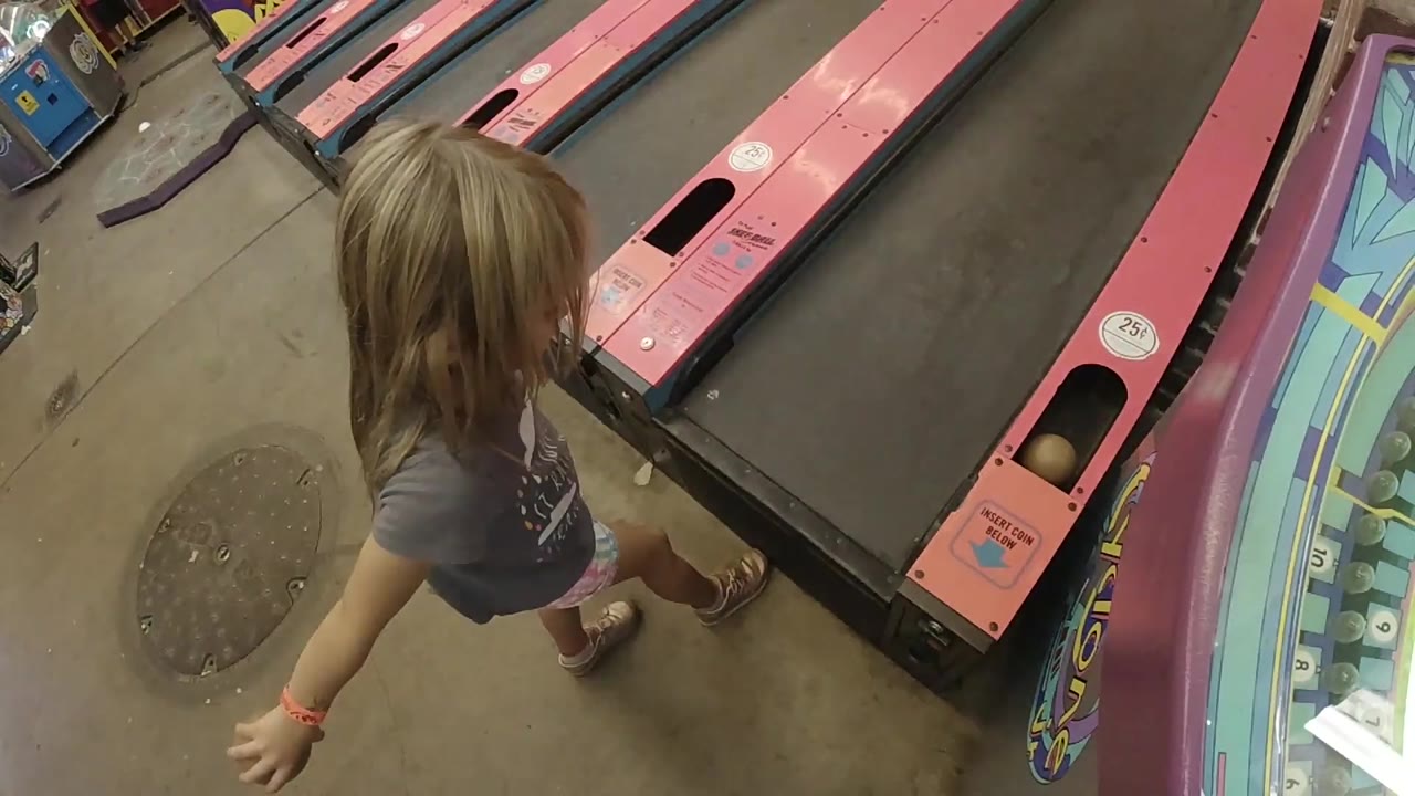 A young blonde girl in Colorado stands close to the ball return opening on a pink skee ball lane, with colorful arcade trim along the right edge.