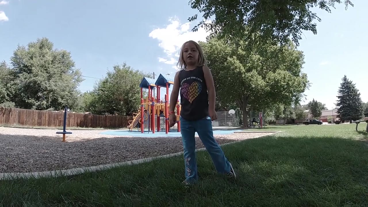 The child remains standing under tree shade in the Colorado park while the colorful play structure and pale blue sky fill the background.