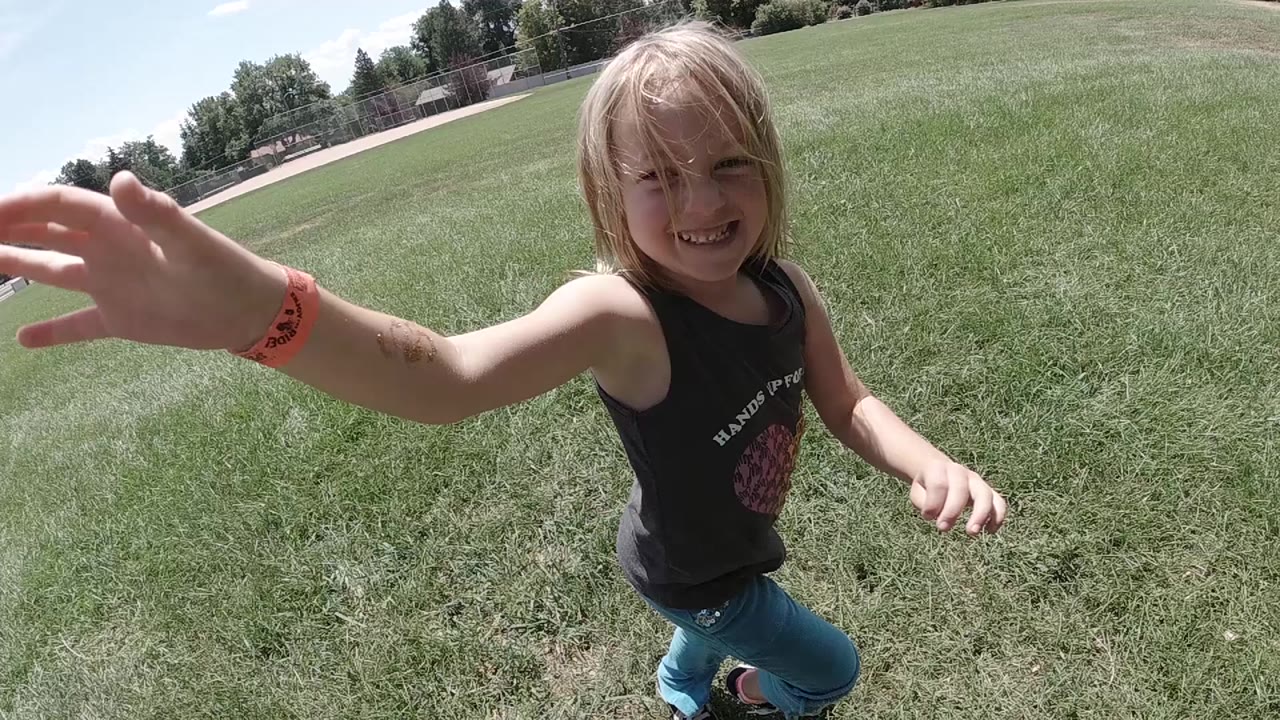 A smiling blonde young girl in Colorado reaches one hand toward the wide camera while standing on bright grass.