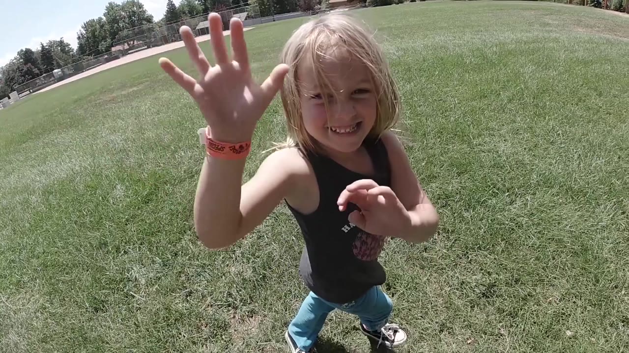 The young girl smiles broadly in Colorado with both hands raised near the lens and a wide lawn behind her.