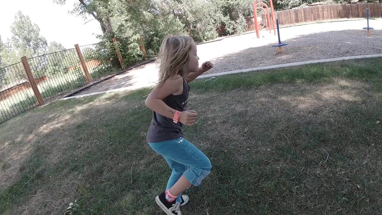 A young girl runs across shaded grass in a Colorado park, with orange playground equipment and a wooden fence in the background.