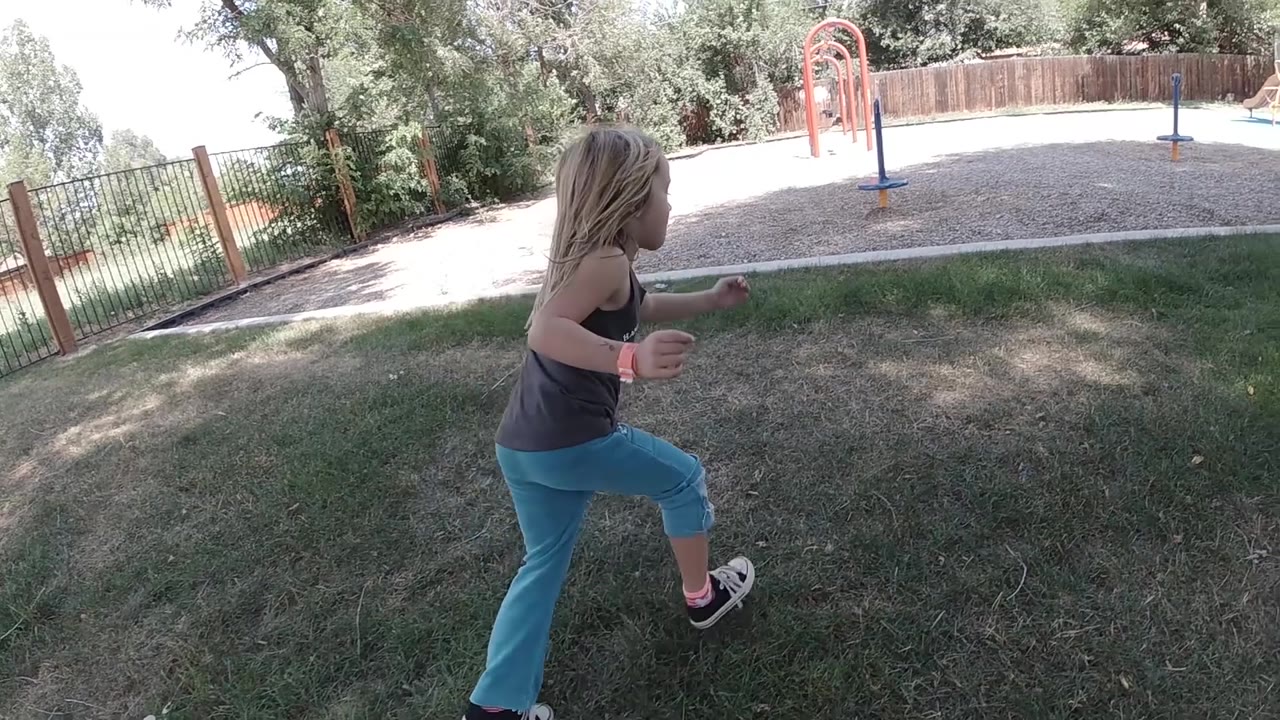 A young girl lifts her knee mid-stride on the grass, with shaded trees on the left and playground structures beyond the gravel area.