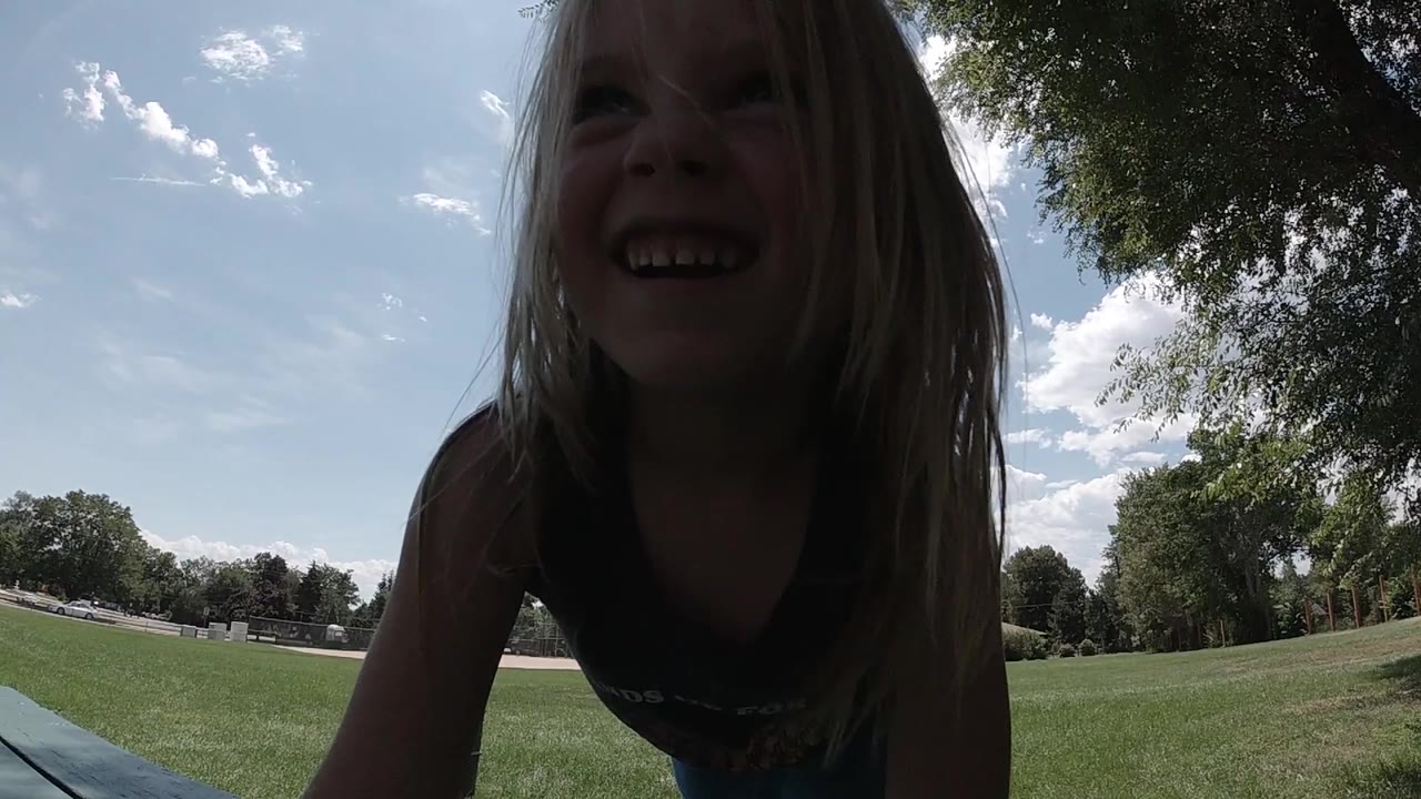 A young blonde girl laughs very close to a low camera in a Colorado park, with bright sky, grass, and trees behind her.