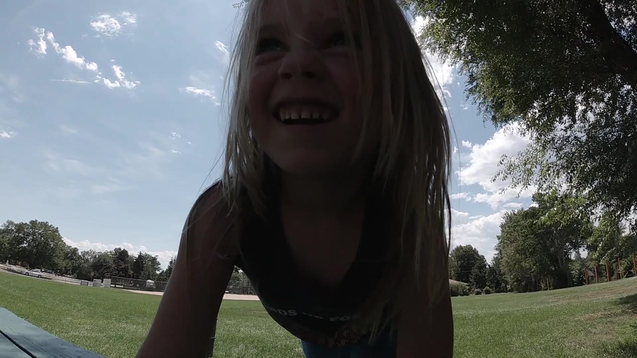 The girl remains close to the lens in Colorado, smiling in shadow against a bright lawn, blue sky, and tree line.